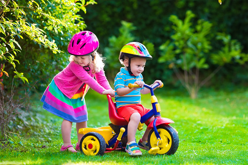 Kids riding bikes in a park. Children enjoy bike ride in the garden. Girl on a bicycle and little boy on a tricycle in safety helmet playing together outdoors. Preschool child and toddler kid biking.