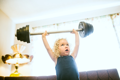A toddler aged girl holds a toy barbell over her head in a cross training style snatch and grab, or military press.  She stands in her living room, bright light shining in the windows behind her.  Horizontal image with copy space.