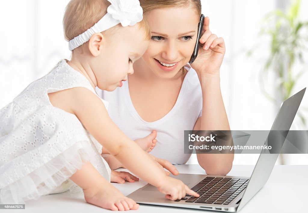 mother and baby daughter works with  computer, phone young mother with baby daughter works on the Internet with a computer and phone Computer Stock Photo