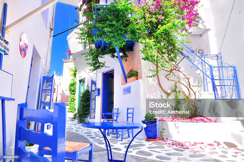 Narrow whitewashed street of Skiathos, Greece Traditional greek architecture in a narrow alley in Skiathos town, Sporades archipelago, Greece with cobblestone pathways, blue painted windows, door and furniture, whitewashed walls and beautiful bougainvillea in full bloom. All signs are edited except the one that says "to the old port (palio limani)" which shows the way to the old port of Skiathos. This shot has a low vantage point for giving the dramatic feeling of entering the picture. Skiathos Stock Photo