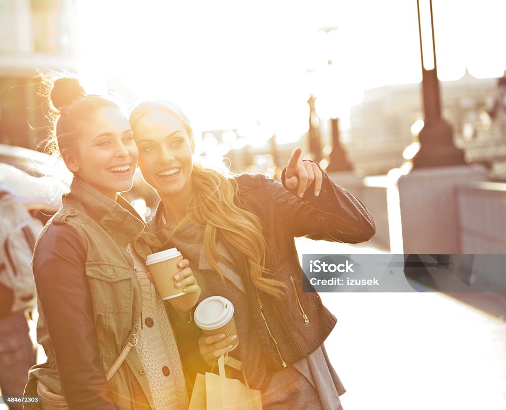Outdoor photo of two women tourists in London Two young women in London.  Each woman is carrying a paper coffee cup.  One woman is pointing at something and smiling, and the other woman is looking in that direction and smiling as well.  The sun is casting a light upon them. Coffee - Drink Stock Photo