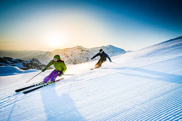 Man and woman skiing downhill at dusk, snowcapped mountain in background.