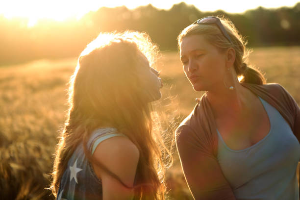 Mother and daughter standing in a ripe barley field in summertime. Their mouthes are ready to kiss each other. The teenage girl with long blond hair is wearing a blue jeans dress printed with white stars. The mother has long blond hair in a ponytail. She is wearing a light blue shirt, a brown bolero jacket, and sunglasses. Both having a caucasian ethnicity and a northern european descent. Taken with selective focus from front view and side view, back lit by the sun with some lens flares. Sunbeams, field and trees in the background. Taken in Sande, Friesland, Lower Saxony, Germany, Europe.