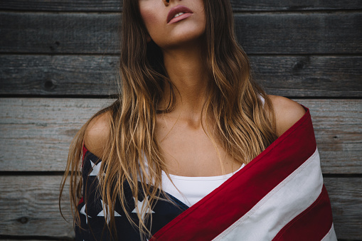 Young woman wrapped in American flag standing outside by wooden wall. Caucasian, long blond hair. Copy space.