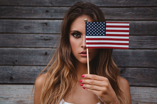 Young woman holding American flag standing outside by wooden wall. Caucasian, long blond hair.