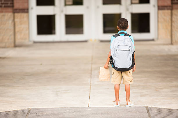 African descent, little boy faces school building. Lunch, backpack. Back to school... African descent little boy ready for the first day of school. The student holds his lunch sack and carries a backpack. The elementary school building entrance is in the background.  Rear view.  sad child standing stock pictures, royalty-free photos & images