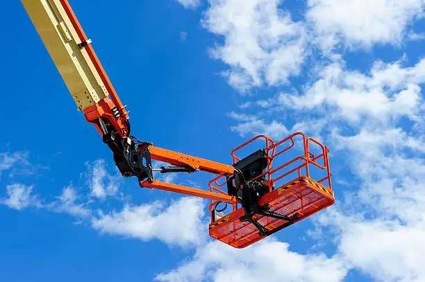 Cherry picker work bucket platform and hydraulic construction cradle of lifting arm painted in orange and beige colors with white clouds and blue sky on background, heavy industry machinery vehicle 