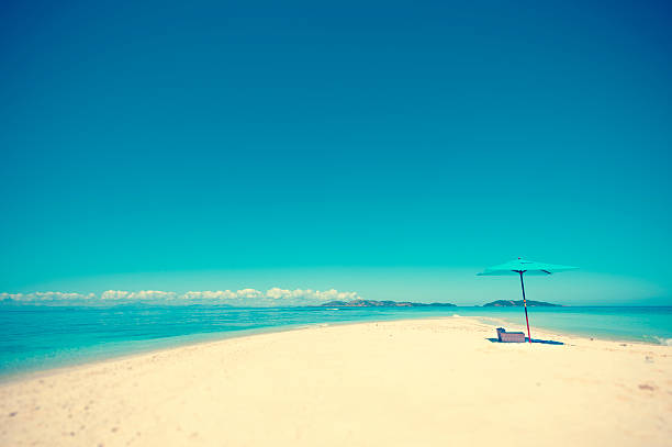 Beach umbrella with picnic on a remote sand island beach. The Fiji beach is deserted with turquoise blue water and pure white sand. This particular sandbar is only visible at low tide. Very romantic getaway. Wide angle shot showing most of the island. Cross processed. Copy space