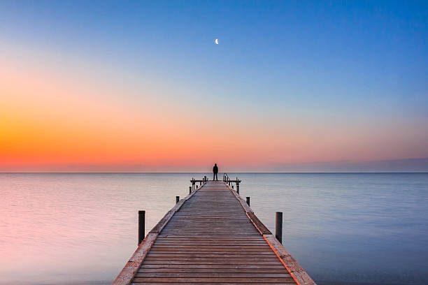 Man standing on jetty at beach with sunrise and moon A man standing at the end of a jetty watching the moon in the sky, at sunrise sunset time. Silhouette man. Beach landscape sea bridge stock pictures, royalty-free photos & images