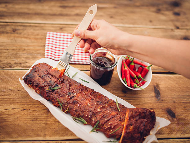 Person's hand painting a basting onto a tasty looking rack of ribs sprinkled with fresh rosemary on a wooden table with a jar of barbecue sauce and fresh red chillies