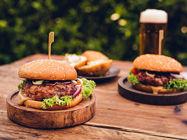 Huge gourmet cheese burgers on a rustic wooden table outdoors Delicious home made gourmet cheese burgers made from healthy prime beef with fresh ingredients placed on wooden platters on a rustic wooden table with a tall glass of beer in the background outdoors at a back yard barbecue grill burgers stock pictures, royalty-free photos & images
