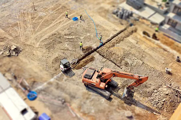 Manual workers and Engineers are digging a raw in the ground, they do drainage, sewer system, then make a new asphalt road. Directly above, tilt-shift.