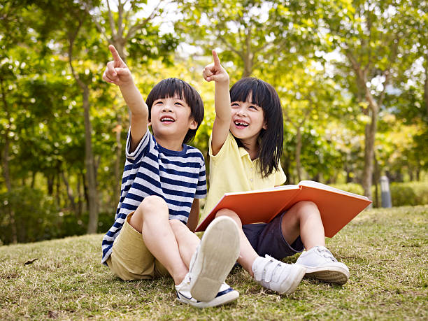 two beautiful asian children little asian boy and girl with book in hand sitting on grass pointing to the sky. japanese boy and girl stock pictures, royalty-free photos & images