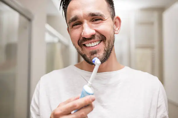 Handsome latin man holding an electric toothbrush and smiling. He has black hair, beard and he is wearing white pajamas. Bathroom interior is on the background.