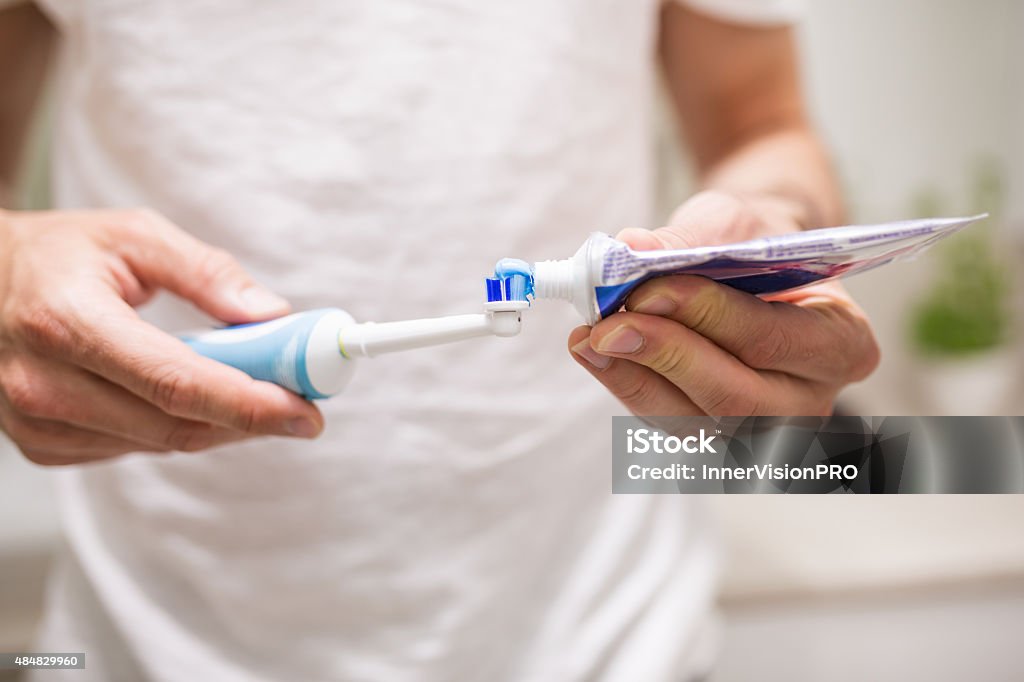 Always use gum protective toothpaste Close up of human hands pouring blue toothpaste on an electric toothbrush. Man is blurred, in a white t-shirt. There's bathroom visible on the background Electric Toothbrush Stock Photo