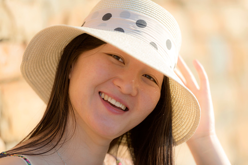 Smiling young female Chinese with long dark hair, wearing a white straw hat with polkadots ribbon and sleeveless shirt.  Koper, Slovenia, Adriatic Sea, Europe. Sandy brown colour. Stone wall as background. Nikon D800, full frame, XXXL.