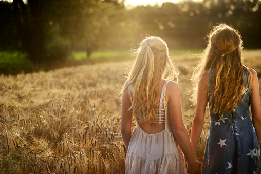 Two teenage girls with long blond hair standing in a ripe barley field in summertime. They are holding hands. One girl is wearing a white sundress, the other one is wearing a blue jeans dress printed with white stars. Both girls have a caucasian ethnicity and a northern european descent. Taken with selective focus from rear view, back lit by the sun. Sunbeams, field and trees in the background.