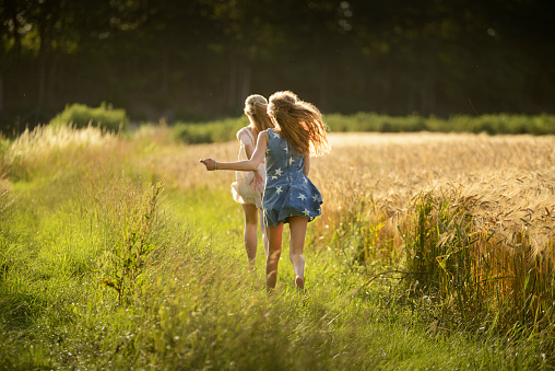 Two teenage girls with long blond hair running on a green meadow at a ripe barley field in summertime. One girl is wearing a white sundress, the other one is wearing a blue jeans dress printed with white stars. Both girls have a caucasian ethnicity and a northern european descent. Taken with selective focus from rear view, back lit by the sun. Field and trees in the background.