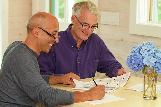Senior Gay Male Couple Working Together on Financial Documents Senior gay male couple, smiling and affectionate, working at their kitchen table on financial documents such as wills, financial planning, or a contract for a house. gay dinner table stock pictures, royalty-free photos & images
