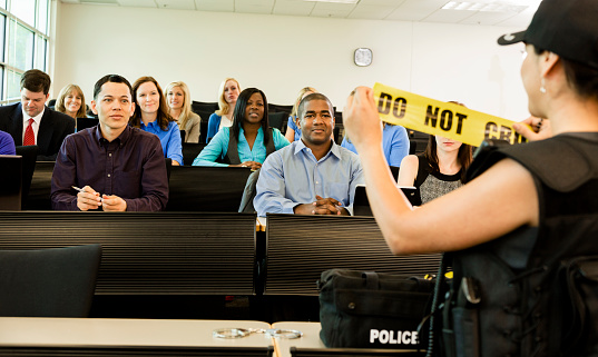 Latin descent policewoman speaks to potential police cadets in a lecture hall;  OR  policewoman gives a safety presentation to local community members.  She wears her police uniform and holds cordon tape. Large classroom setting. 