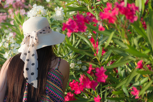 Portrait of a pretty young female Chinese with long dark hair, wearing a white straw hat with polkadots ribbon and sleeveless shirt. She is smelling blossoms  of white and red  rhododendron bushes. Rear view. Koper, Slovenia, Europe. Nikon D800, full frame, XXXL.