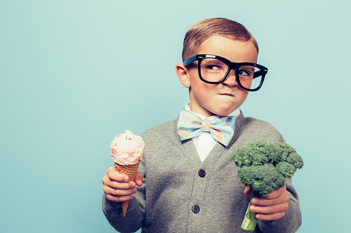 A young nerd boy with glasses is not sure whether to eat the ice cream cone or the broccoli. He is not sure which one is healthier.  