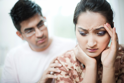 Indoor shoot of depressed couple, young woman who is holding her head out of headache, depression or work stress may be due to relationship or other problems while her young male partner consoling her by giving gentle touch on her shoulders from behind. The woman is in shirt and her friend is in t-shirt. Horizontal, head and shoulders composition with selective focus.