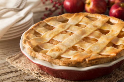 A lattice apple pie.  The apple pie is set in a fall scene with apples, dessert plates, forks, and seasonal decoration.  The pie is set on a rustic farm table with burlap textured hot pad.  This image could be used to illustrate:  Baking, Fall, Autumn, Thanksgiving, Apple pie, holiday desserts, holiday recipes, etc