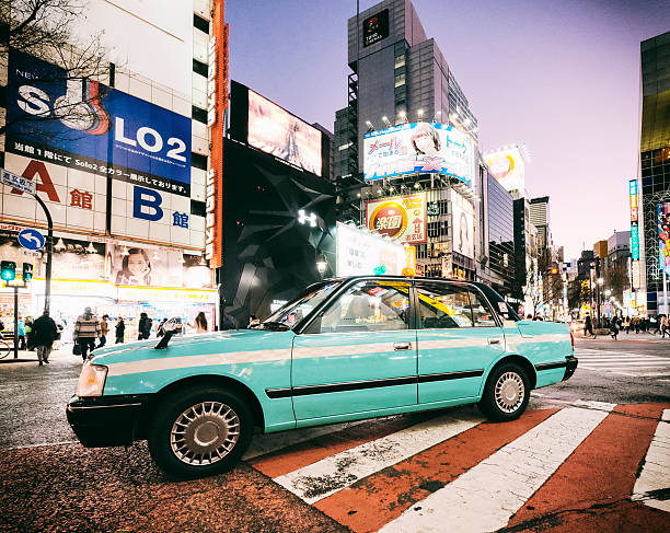 A nighttime street scene in the Shibuya area of Tokyo, Japan. A taxi sits on a zebra crossing, waiting for passengers.