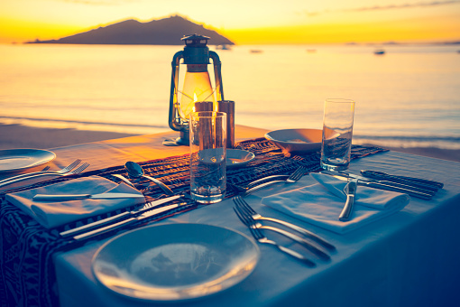 A table for two at a beach restaurant. The table is right on the beach at sunset with a lovely view of the water. Table is set for several courses with white table cloth. Absolute waterfront with lapping waver. Luxurious setting in a tranquil, romantic and remote location. Cross processed with copy space.