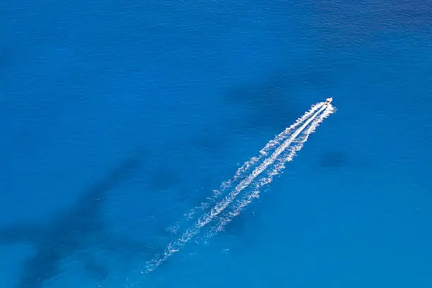 High view aerial image of powerboat floating in a turquoise blue sea water. The boat is moving diagonally through the frame of the image.
