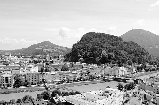 Salzach river flows through Salzburg city centre in Austria. Pedestrians cross the Makartsteg bridge. The Kapuzinerberg rises above the city, with the Kuehberg and Gaisberg mountains beyond - monochrome processing.