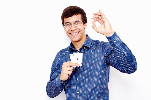 Young hispanic man wearing blue shirt and glasses holding two aces (clubs and hearts) in his hand and showing A-ok hand gesture with smile against white wall - gambling concept