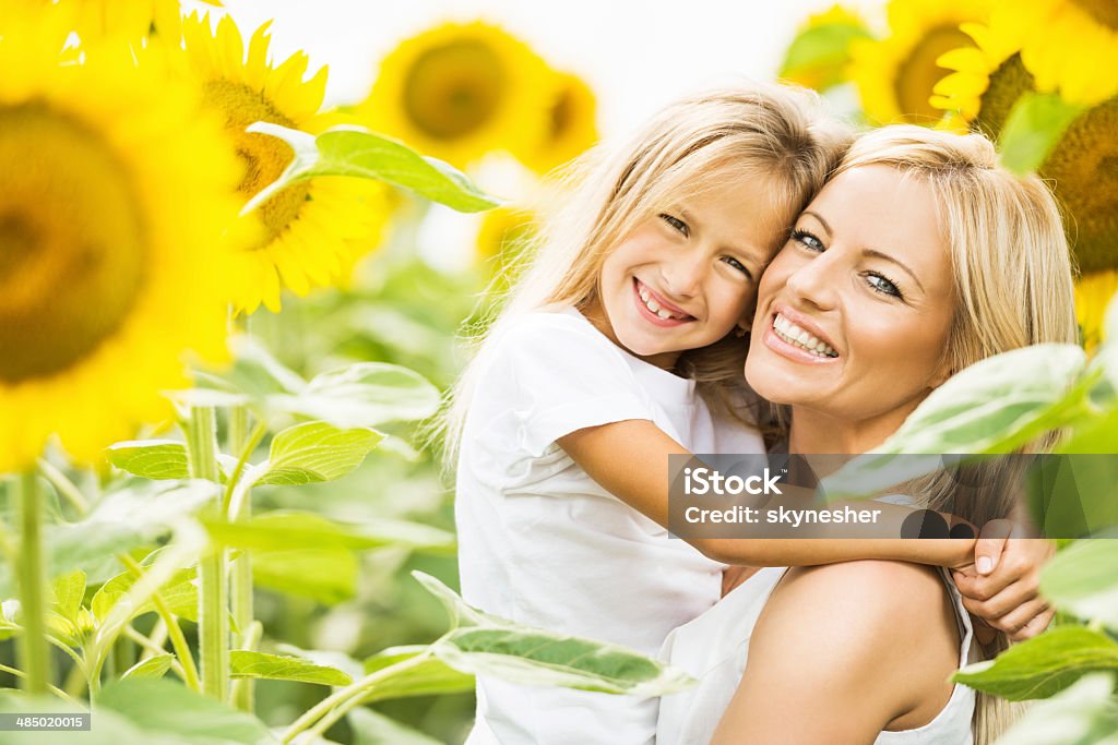Smiling mother and daughter among sunflowers. Happy mid adult mother holding her little daughter among sunflowers and looking at the camera.    Mother Stock Photo