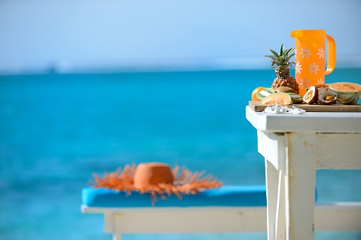Freshly cut Mauritian fruit and an orange jug offset the beautiful turquoise sea in Mauritius. An orange hat lies on a blue cushion and bench in the background. Copy space allowed.
