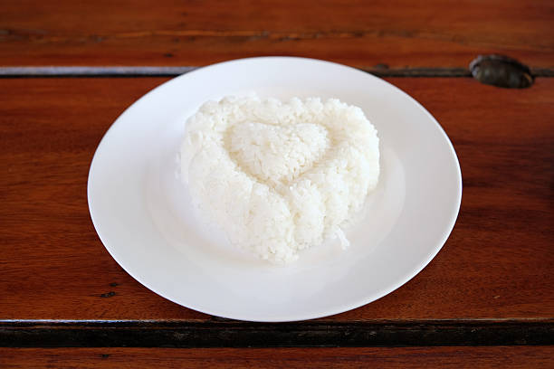 A beautifully plated serving of steamed jasmine rice in a traditional blue and white Thai ceramic bowl, set on a red decorative surface with floral accents. The clean white background enhances the simplicity and elegance of the composition, highlighting the cultural significance of Thai cuisine. The contrast between the blue and red elements creates a visually appealing representation of classic Asian dining aesthetics, perfect for showcasing authentic meals and traditional tableware.