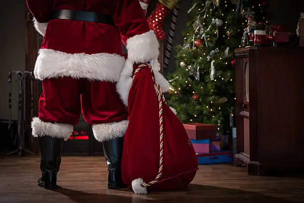 Real authentic Christmas photo of Santa Claus from behind and waist down, holding his bag and standing in a living room in front of the fireplace.