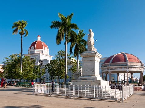 Cienfuegos, Cuba - March 20, 2010: Monument to Josi Marti, surrounded by metal railings, in the  Plaza de Amaris, with people and two red domed buildings of Colonial Architecture in the background