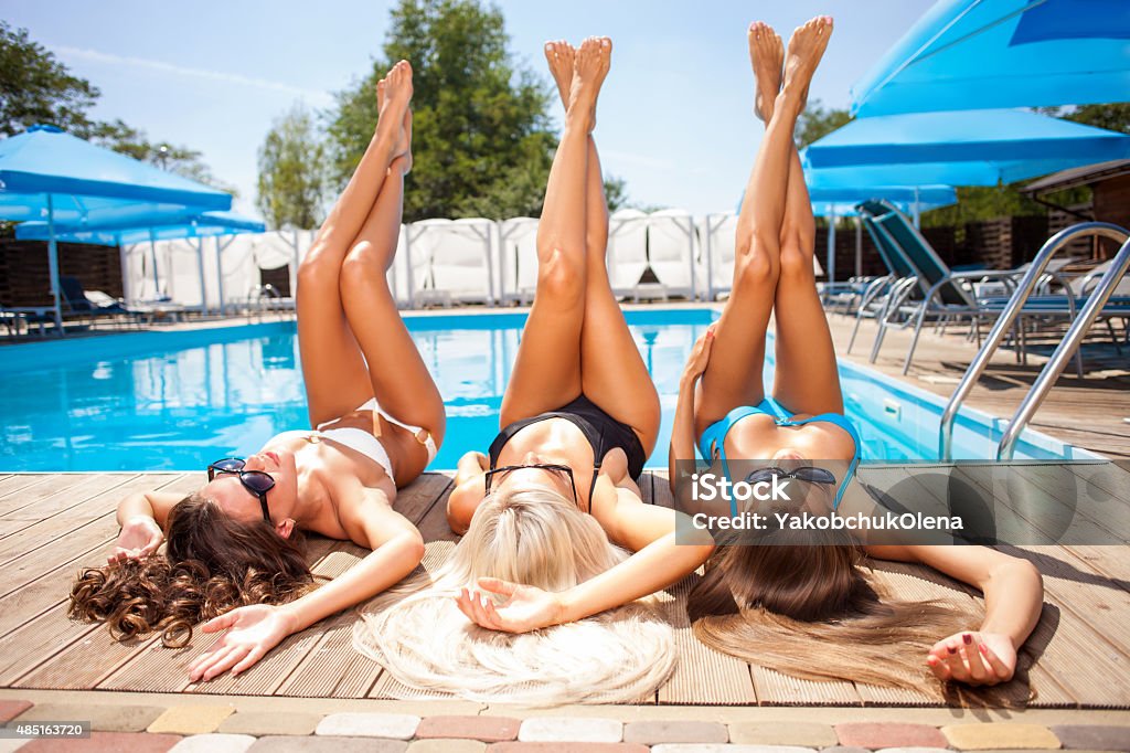 Cheerful young girls are getting a suntan on holidays Attractive women are lying near a swimming pool and relaxing. They are sunbathing with pleasure. The friends are raising their legs up 2015 Stock Photo