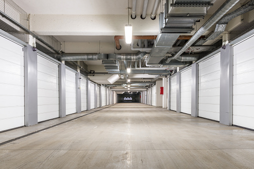 An underground parking structure sits empty, its beige floor stretching out toward a dark exit ahead.  On either side are long rows of closed storage bays with white doors.  The bays are numbered.  The ceiling is also beige and is crisscrossed with pipes and ducts of various sizes.  Light fixtures are spaced intermittently overhead.