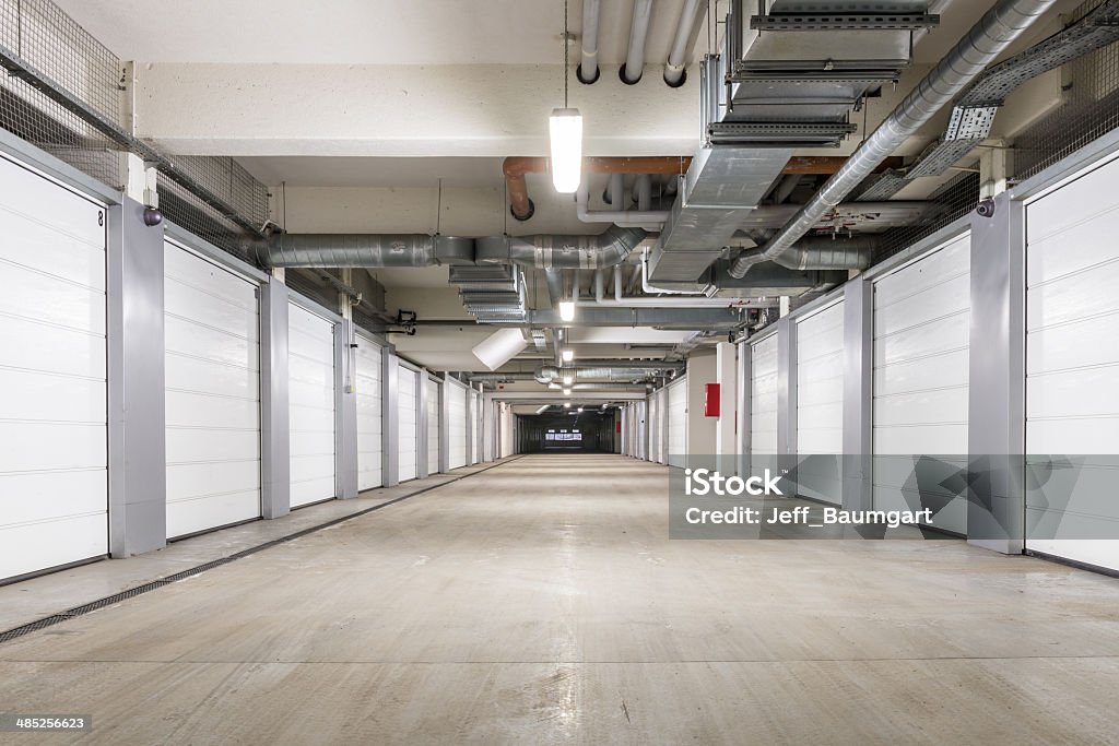 Interior of underground parking garage in Europe An underground parking structure sits empty, its beige floor stretching out toward a dark exit ahead.  On either side are long rows of closed storage bays with white doors.  The bays are numbered.  The ceiling is also beige and is crisscrossed with pipes and ducts of various sizes.  Light fixtures are spaced intermittently overhead. Storage Compartment Stock Photo