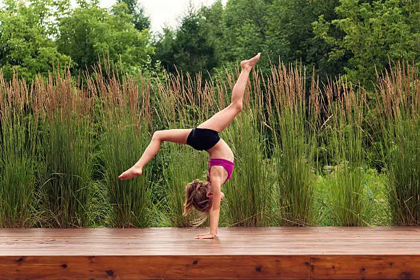 Your kid is full of energy? Maybe she's a future gymnast, like this young lady, who spent most of her days perfecting her moves. She is standing on her hands on a wooden floor outdoors in summer nature. Full length horizontal shot with copy space.