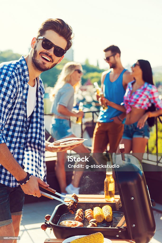 Tasty food and good company. Happy young man barbecuing and looking at camera while three people having fun in the background Barbecue Grill Stock Photo