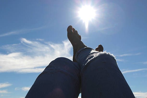 Looking up past woman's feet and ripped jeans to sun, clouds and blue sky