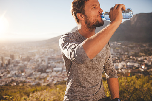 Young runner taking a break after morning run drinking water. Young man relaxing after a running training session outdoors in countryside on sunny day.