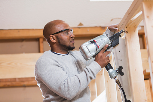 A construction worker, an African American man in his 40s, working on a home remodeling project.  He is standing on a ladder with a nail gun, nailing wood posts.  He is serious, wearing safety glasses.