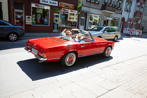 Essen, Germany - August 2, 2015: Capture of caucasian senior couple with straw hats in red Mercedes SL 280 convertible car. In background are cars and buildings. In one building is lottery and tabacco shop.