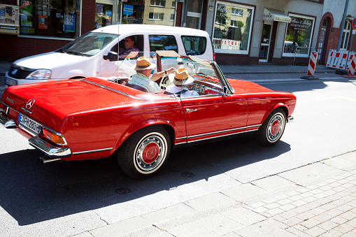 Essen, Germany - August 2, 2015: Summer shot of caucasian senior couple sitting with sraw hats in red Mercedes SL 280 converible car. In background a man is driving in car. Behind are buildings.