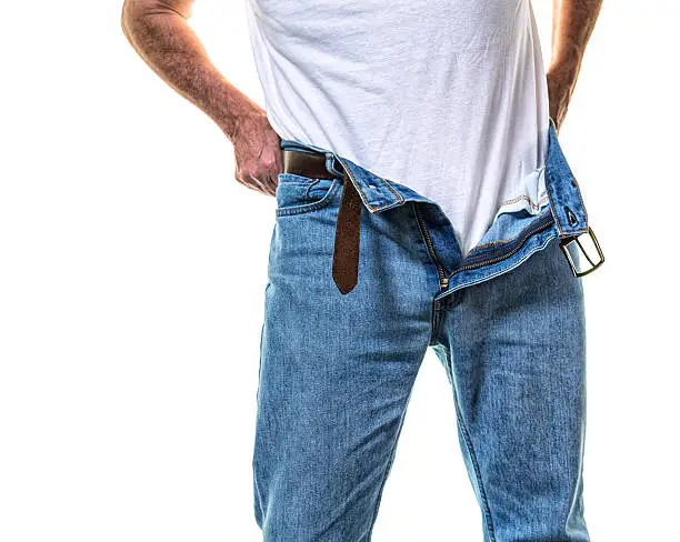 A senior adult man is finishing getting dressed in a white t-shirt and blue jeans. The pants are unzipped and partially open as he adjusts the shirt around his waist.