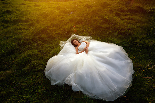 bride lying on grass at sunset, enjoying her wedding day, tranquil scene, summer season.